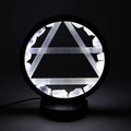 A circular black table lamp with a glowing white geometric triangle design and rough white crystal-like accents, shown on a dark background.