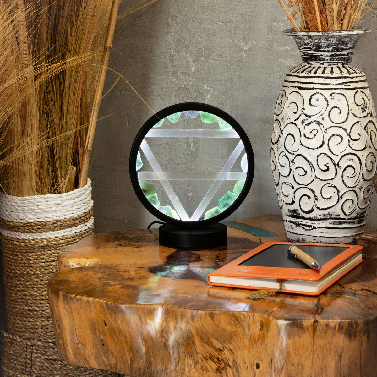 A black circular crystal ball lamp with a triangular prism inside sits on a rustic wooden table, styled with a patterned ceramic vase, a notebook, and dried grass in a home decor setting.