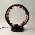 A modern black circular table lamp with a glowing interior lined with polished purple and pink crystal stones, resting on a round black base against a neutral light gray background.