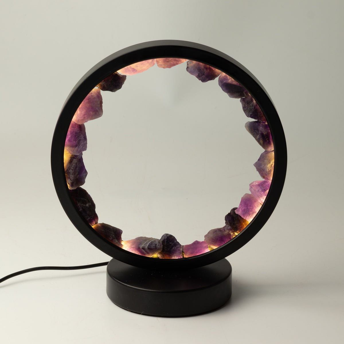A modern black circular table lamp with a glowing interior lined with polished purple and pink crystal stones, resting on a round black base against a neutral light gray background.