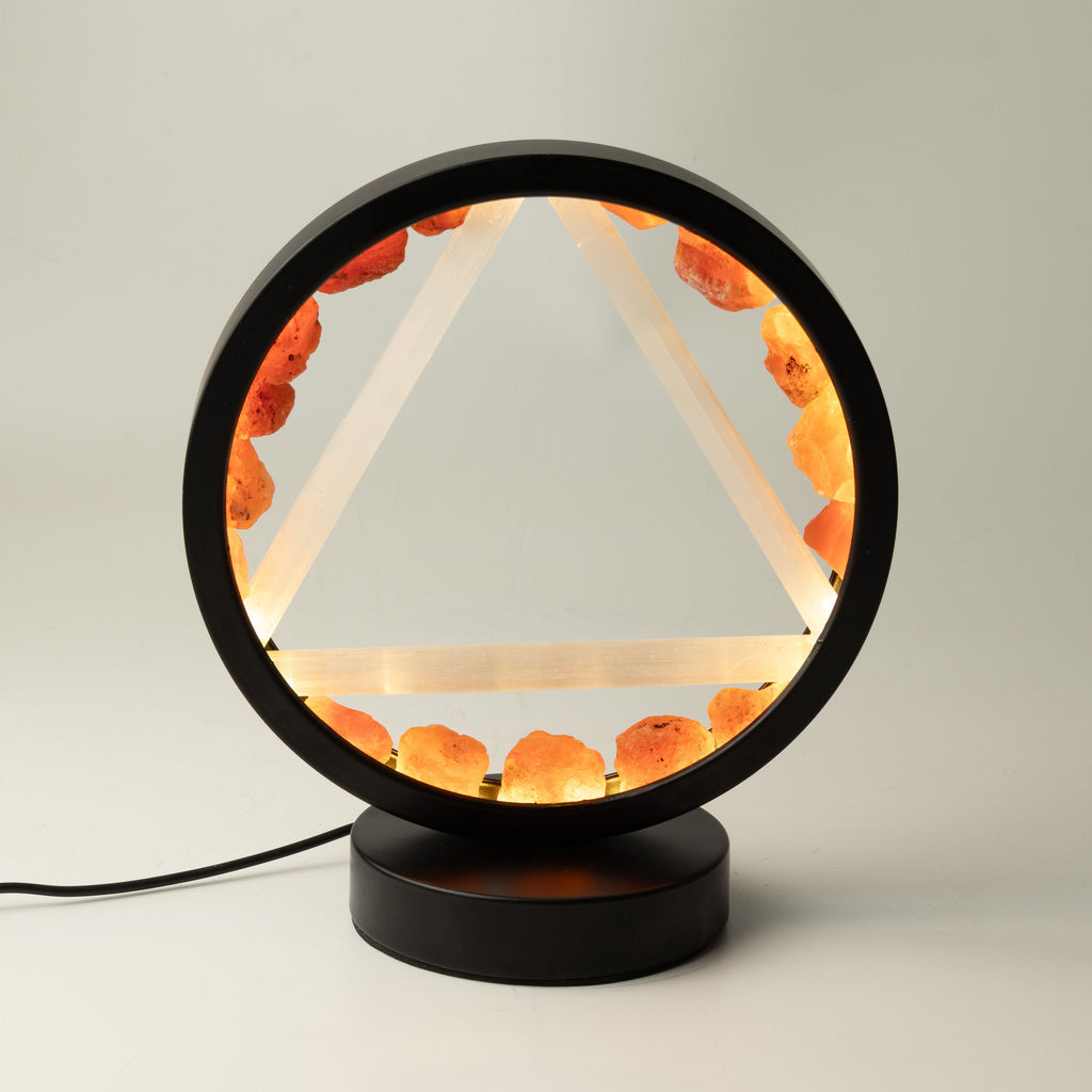 A modern black table lamp featuring a circular frame with a glowing triangular geometric design and warm orange crystal accents, set on a round black base against a neutral light gray background.