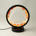 A modern black table lamp featuring a circular frame with a glowing triangular geometric design and warm orange crystal accents, set on a round black base against a neutral light gray background.