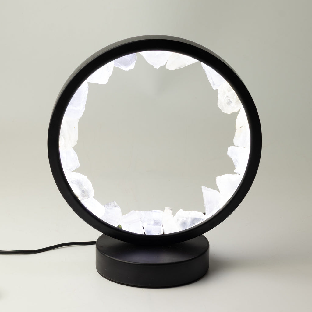 A modern black table lamp with a circular frame and a base, featuring a glowing white interior made of irregular crystal-like shapes, shown on a light gray background.