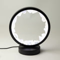 A modern black table lamp with a circular frame and a base, featuring a glowing white interior made of irregular crystal-like shapes, shown on a light gray background.
