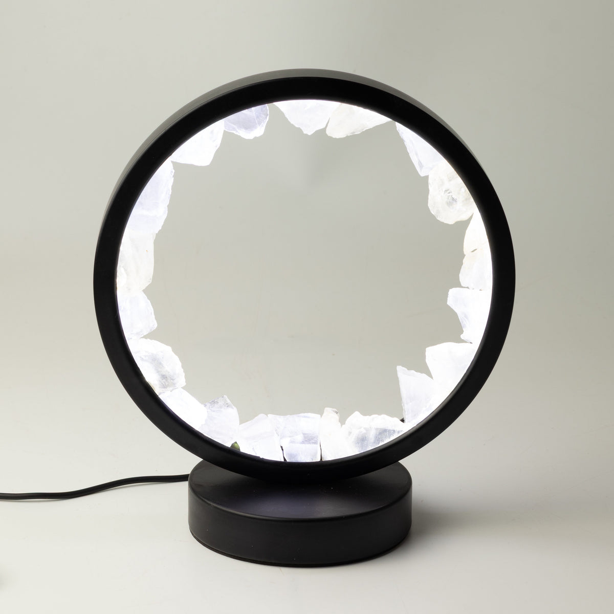 A modern black table lamp with a circular frame and a base, featuring a glowing white interior made of irregular crystal-like shapes, shown on a light gray background.