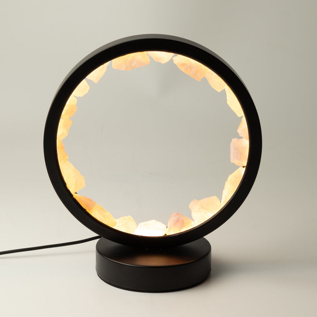 A modern circular table lamp with a black metal frame and base, filled with warm glowing pinkish-orange crystal stones, shown on a light gray background.