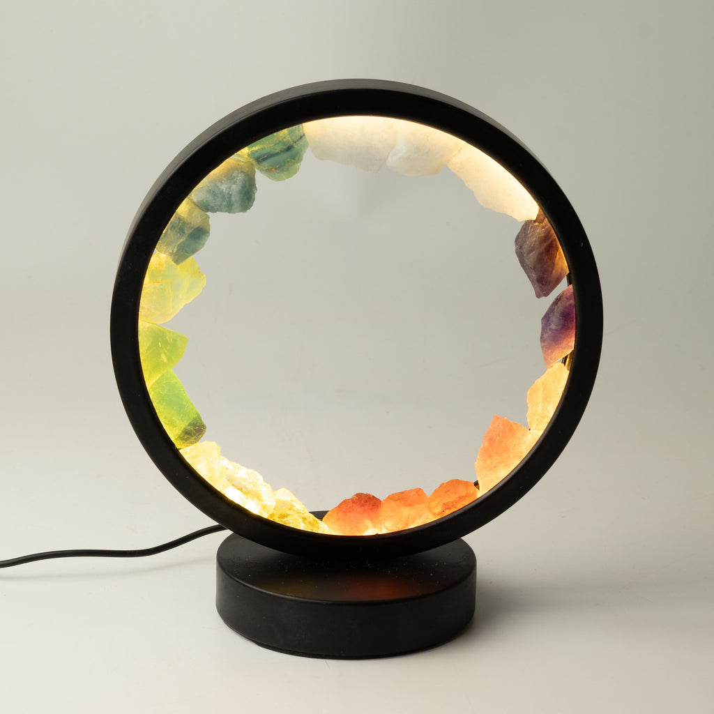 A circular LED table lamp with a black metal frame and base, filled with glowing warm light and set with colorful natural crystal stones in yellow, green, orange, and purple, shown on a neutral light gray background.
