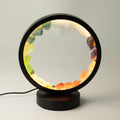 A circular LED table lamp with a black metal frame and base, filled with glowing warm light and set with colorful natural crystal stones in yellow, green, orange, and purple, shown on a neutral light gray background.